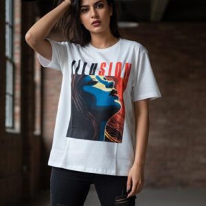 A stylish young woman stands confidently in a modern loft, wearing a white t-shirt featuring bold, colorful artwork of a woman’s profile in blue, red, and yellow tones, with the word “ILLUSION” emblazoned above