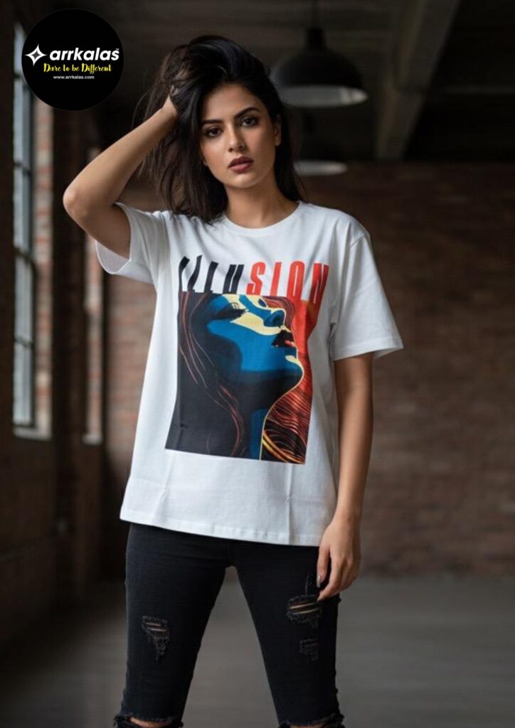 A stylish young woman stands confidently in a modern loft, wearing a white t-shirt featuring bold, colorful artwork of a woman’s profile in blue, red, and yellow tones, with the word “ILLUSION” emblazoned above