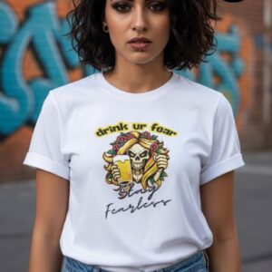 A young woman stands confidently outdoors against a vibrant graffiti wall, wearing a stylish white T-shirt featuring a striking design—a bold cartoon skeleton with flowing golden hair holding a beer, adorned with roses, beneath cheerful yellow text reading "Drink Ur Fear." Below the graphic, elegant black script reads "Stay Fearless," inspiring a rebellious, fearless attitude